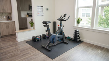 A complete layout setup of an affordable beginner home gym featuring a Wenoker JC302 bike, adjustable weights, and a dense PVC mat in a small apartment corner